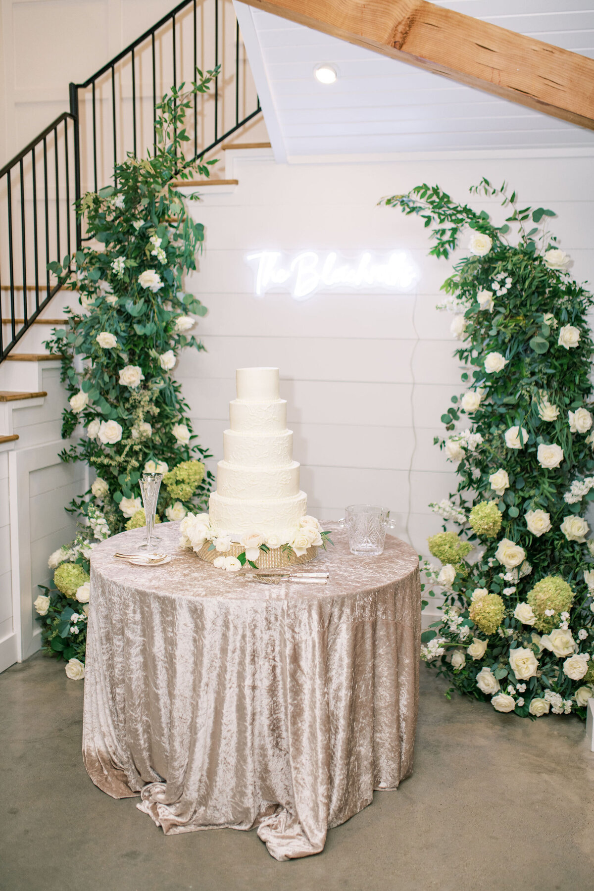 White tiered wedding cake sitting on a table in front of floral arrangements and a neon sign