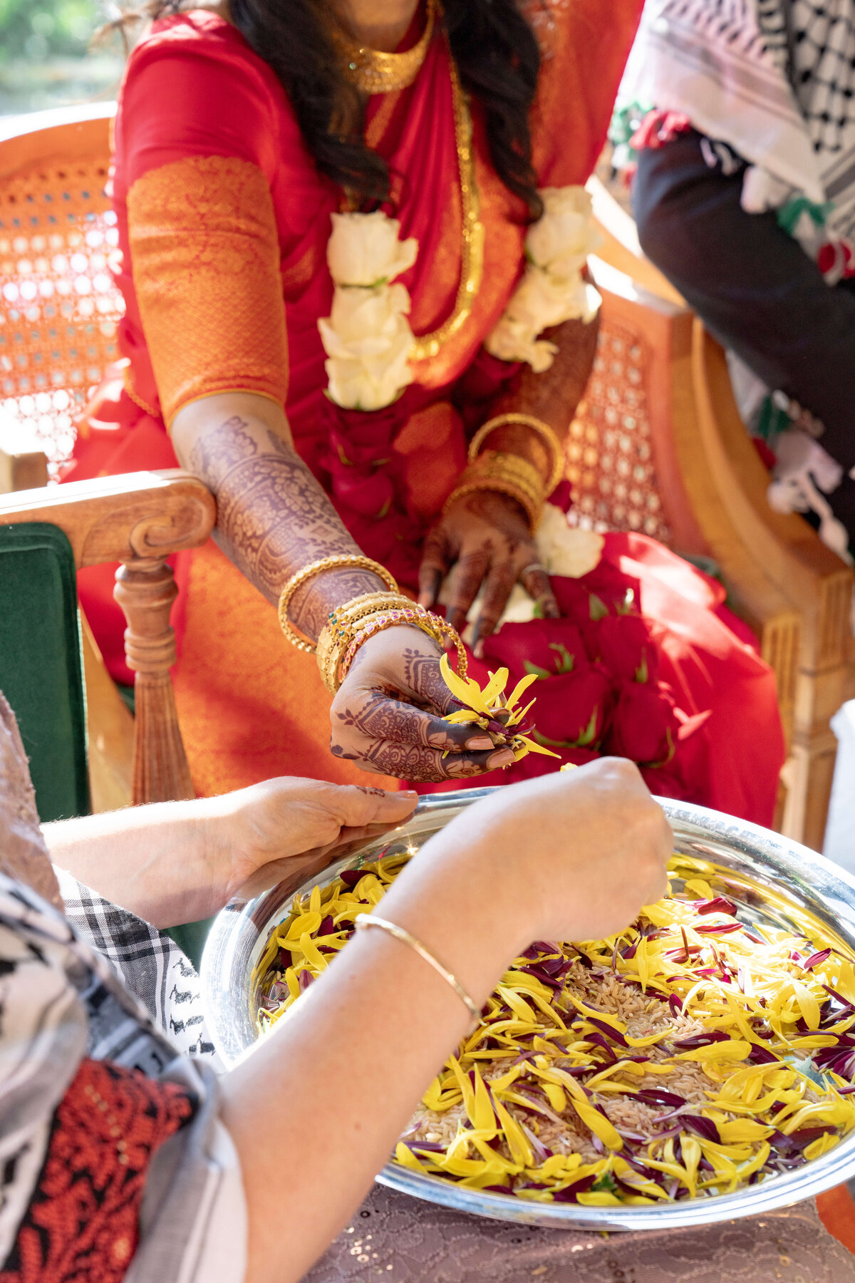 A woman in a red and gold sari and henna-decorated hands receives flower petals from a silver tray held by another person. The setting appears festive, with bright colors and traditional attire.