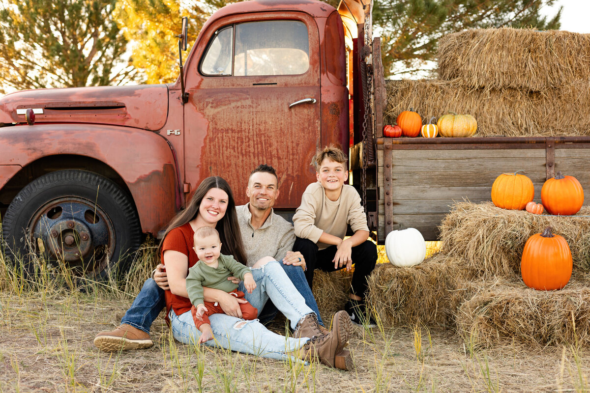 A mom and dad sit in the grass in front of a vintage red farm truck with their pre-teen son and baby boy and they all smile at the camera.