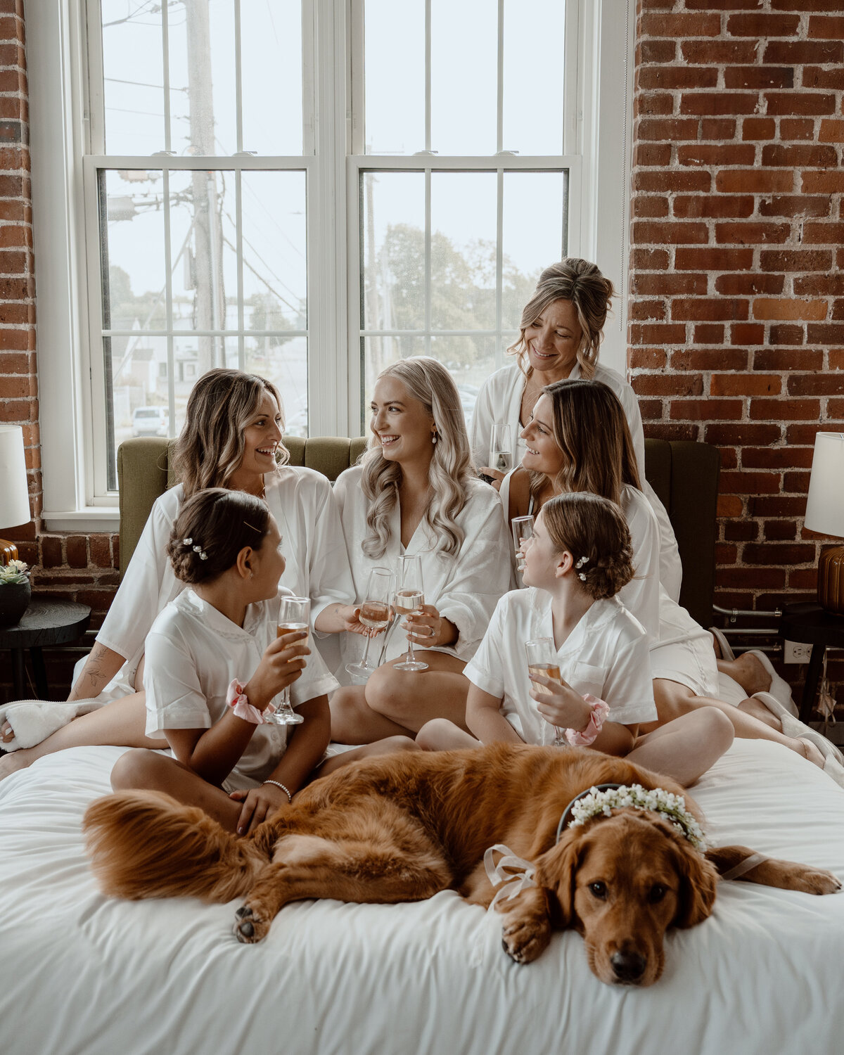 Bride and bridesmaids in matching robes share a joyful moment with champagne glasses, accompanied by a flower-adorned dog, on a cozy bed in a room with brick walls and large windows