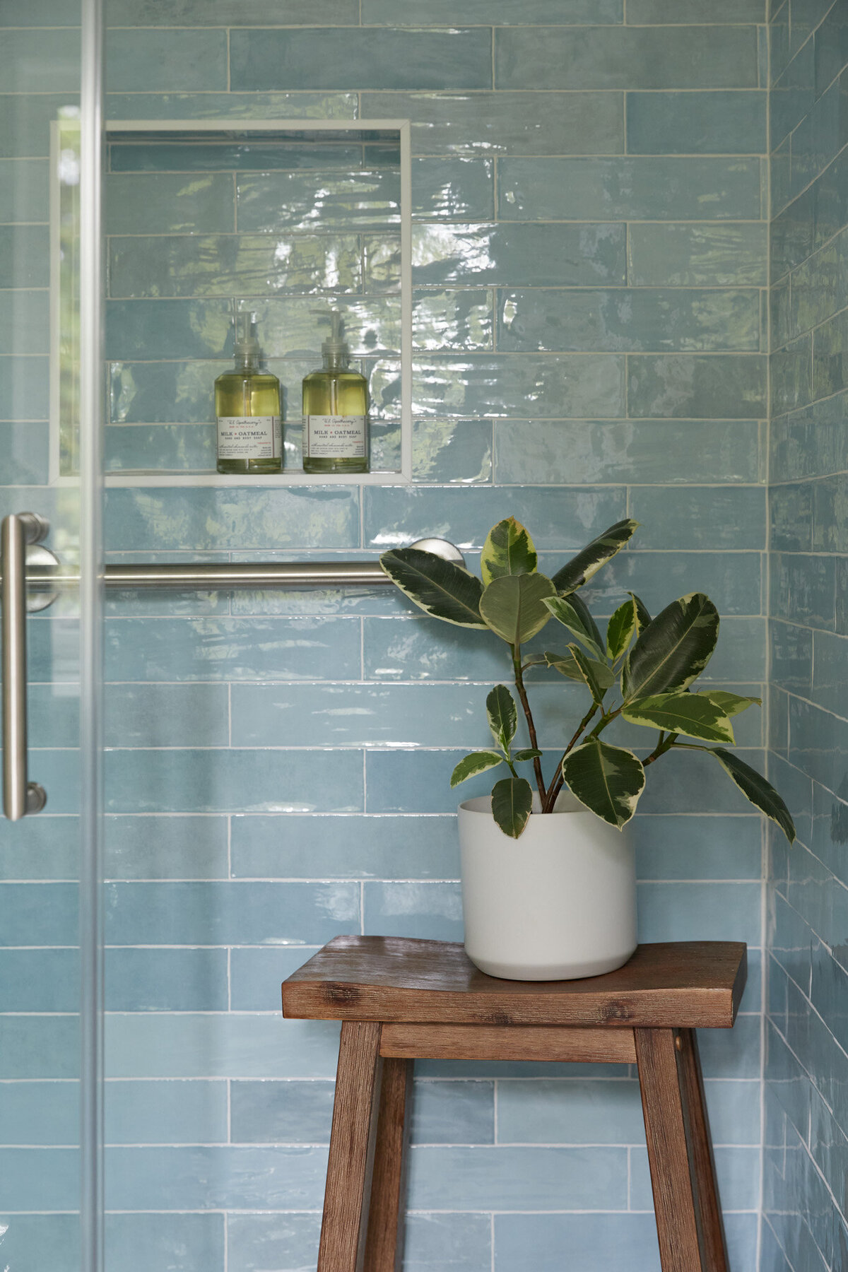 Shower with blue subway tile and a niche with a bottle of soap in it. A wooden stool in the shower with a plant in a white vase sitting on top. White walls to the right of the shower with a silver towel bar and a white towel hanging on it.