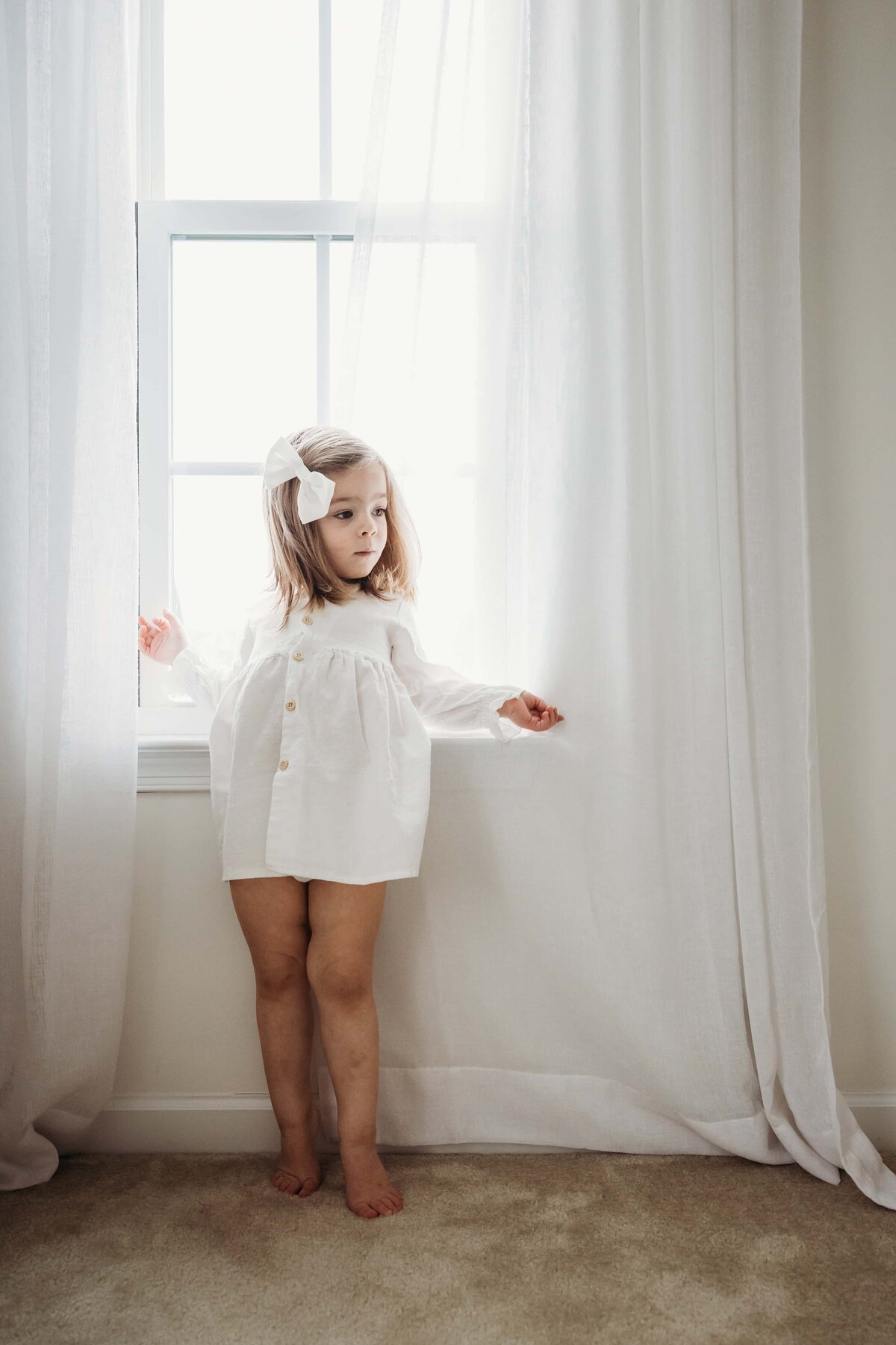 A young child in a white outfit standing between curtains by a window, perfectly captured with lightroom presets for family portraits.