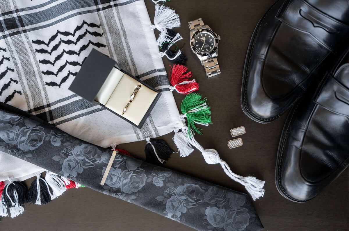 A flat lay of mens accessories on a dark surface: black shoes, a patterned scarf with tassels, a watch, a floral tie, cufflinks, and an open box displaying a wedding band.