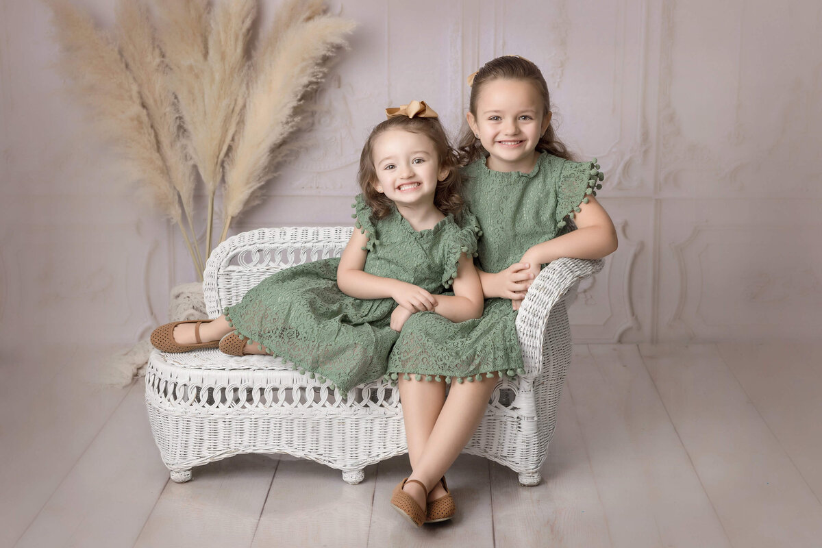 Two young girls sit on a white wicker loveseat, both wearing matching green lace dresses and brown shoes. One girl has a bow in her hair, and both are smiling. The background features tall dried grasses and a light-colored, ornate wall, perfectly captured by the best photographer in Jacksonville.