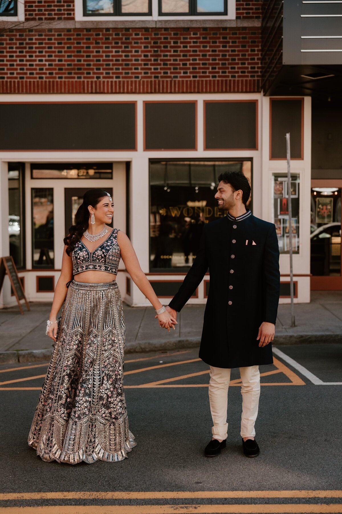 A couple dressed in traditional Indian attire smiles and holds hands in the middle of a street outside Wonderbar in Beacon, NY during a dinner rehearsal. The woman wears a detailed, shimmering lehenga, and the man sports an elegant sherwani with a mandarin collar.