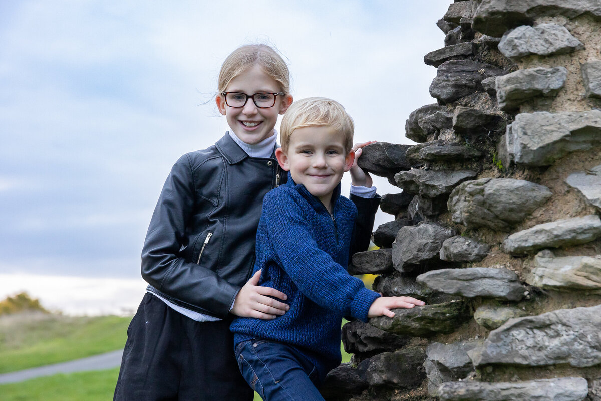 A young girl in a black jacket and glasses smiles while standing behind a younger boy in a blue sweater, both leaning on a stone wall in a grassy area.