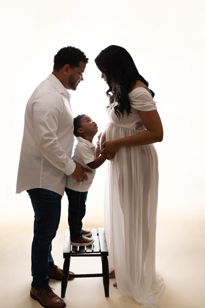 Maternity session with Atlanta, Georgia maternity photographer pregnant woman holding baby bump wearing white sheer matyernity  dress on white backdrop posing with husband and 2 year old son