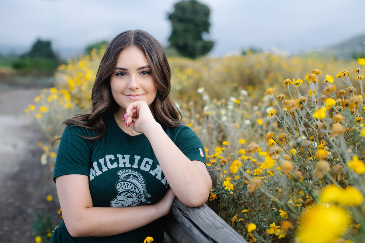 A person with long brown hair, wearing a green Michigan T-shirt, poses with a thoughtful expression near a wooden fence. They are surrounded by a field of yellow flowers under a cloudy sky.