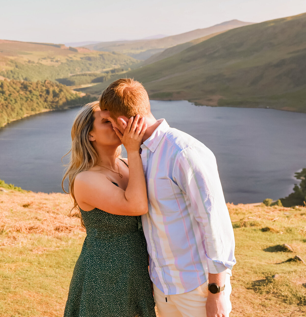 woman wearing green dress and man wearing white and blue striped shirt kissing with mountains in the background