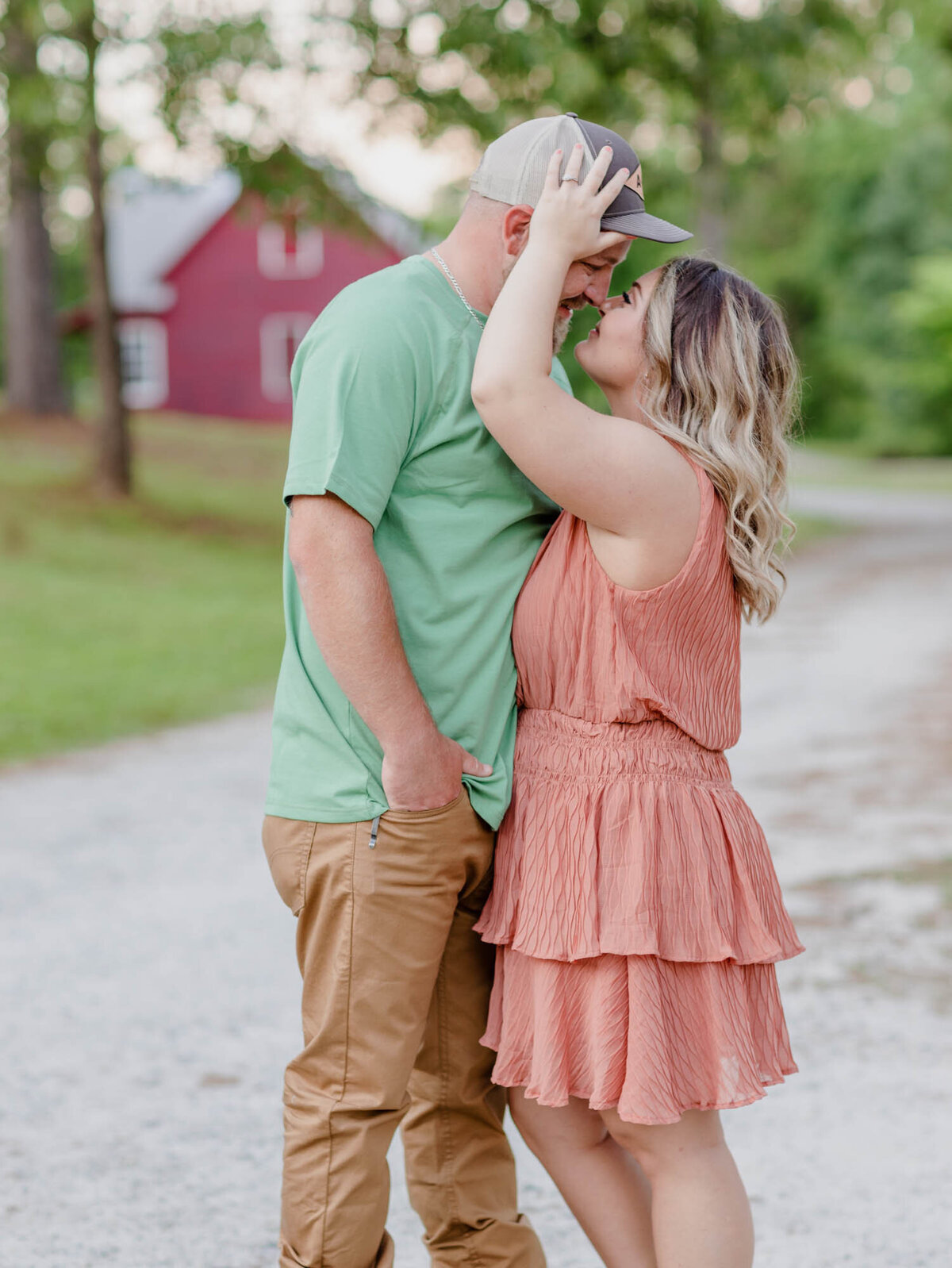 Fun engagement portrait of a woman leaning into him fixing his hat in front of a red barn.