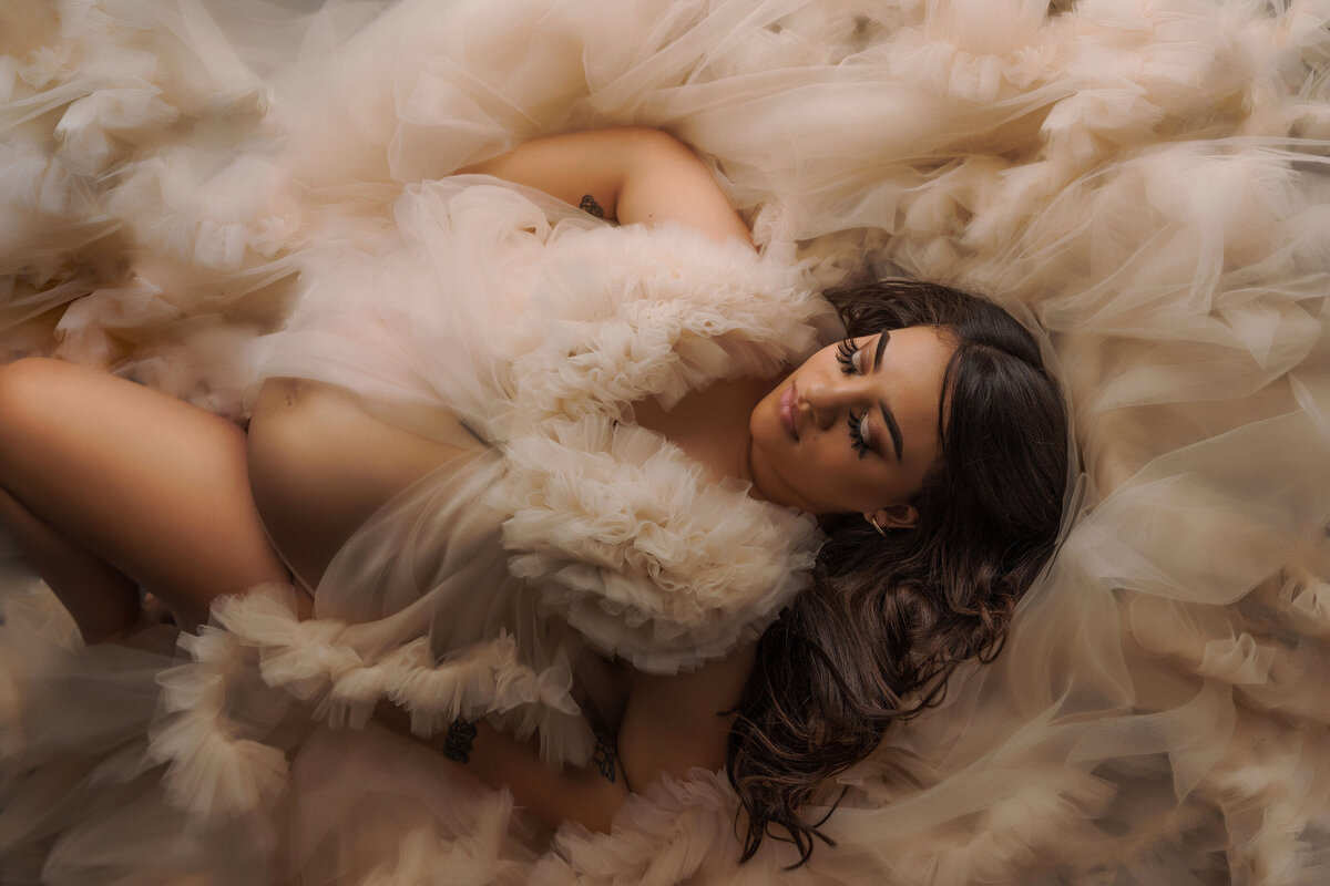 Mother to be laying on her back posed for maternity photo session wearing a fluffy tan gown in studio near dupont