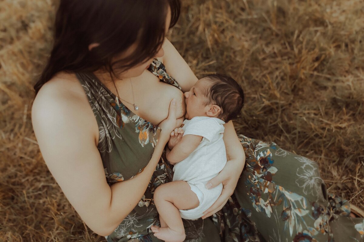 A woman in a floral dress breastfeeds a baby while seated on grass.
