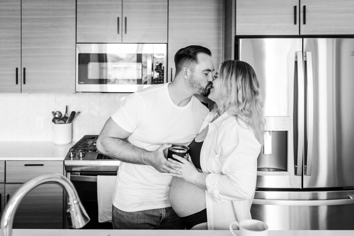 A couple in their kitchen, the woman visibly pregnant, sharing a kiss while holding mugs. The scene is cozy and intimate, with modern kitchen appliances visible in the background.