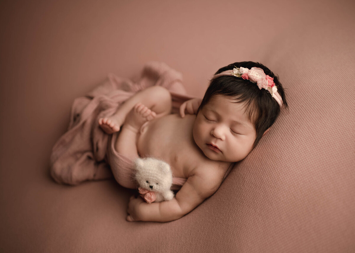 Aerial image. Baby girl is captured for at Lake Elsinore newborn photoshoot. The baby is laing on her back with her legs gently folded atop of her. Her chest is bare and is holding a tiny felt dog under one arm. She is laying on dusty rose stretch fabric and has fabric loosely draped and ruched under her back. Captured by best Lake Elsinore Newborn Photographer Bonny Lynn Photography