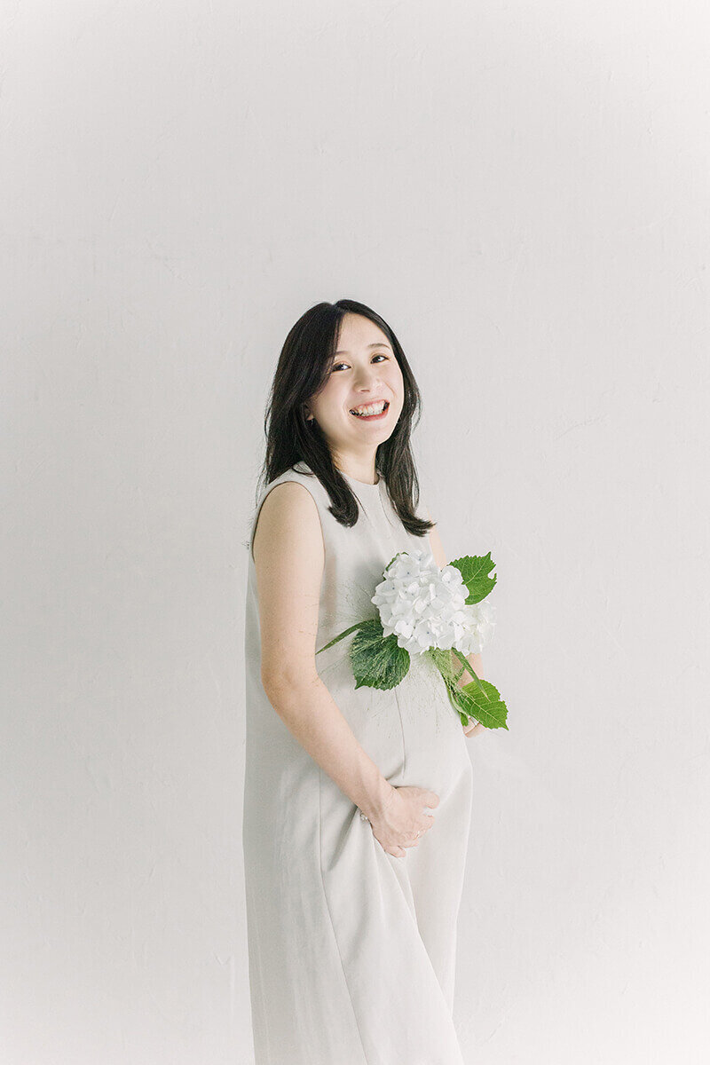 紫陽花を持って笑っている妊婦さんのマタニティフォト。ホワイトコーデで統一された、おしゃれで美しい写真。新宿のスタジオにてYukika Ishikawa Photographyが撮影。