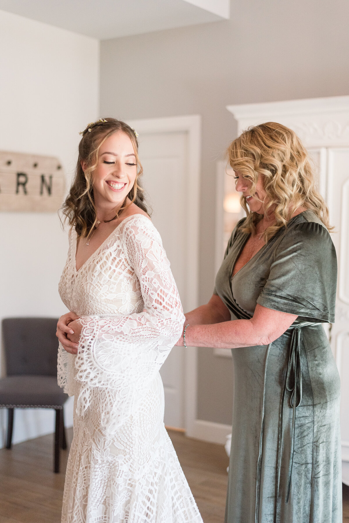 Mom helps her daughter tie up the back of her wedding dress as the bride looks back at her mom smiling