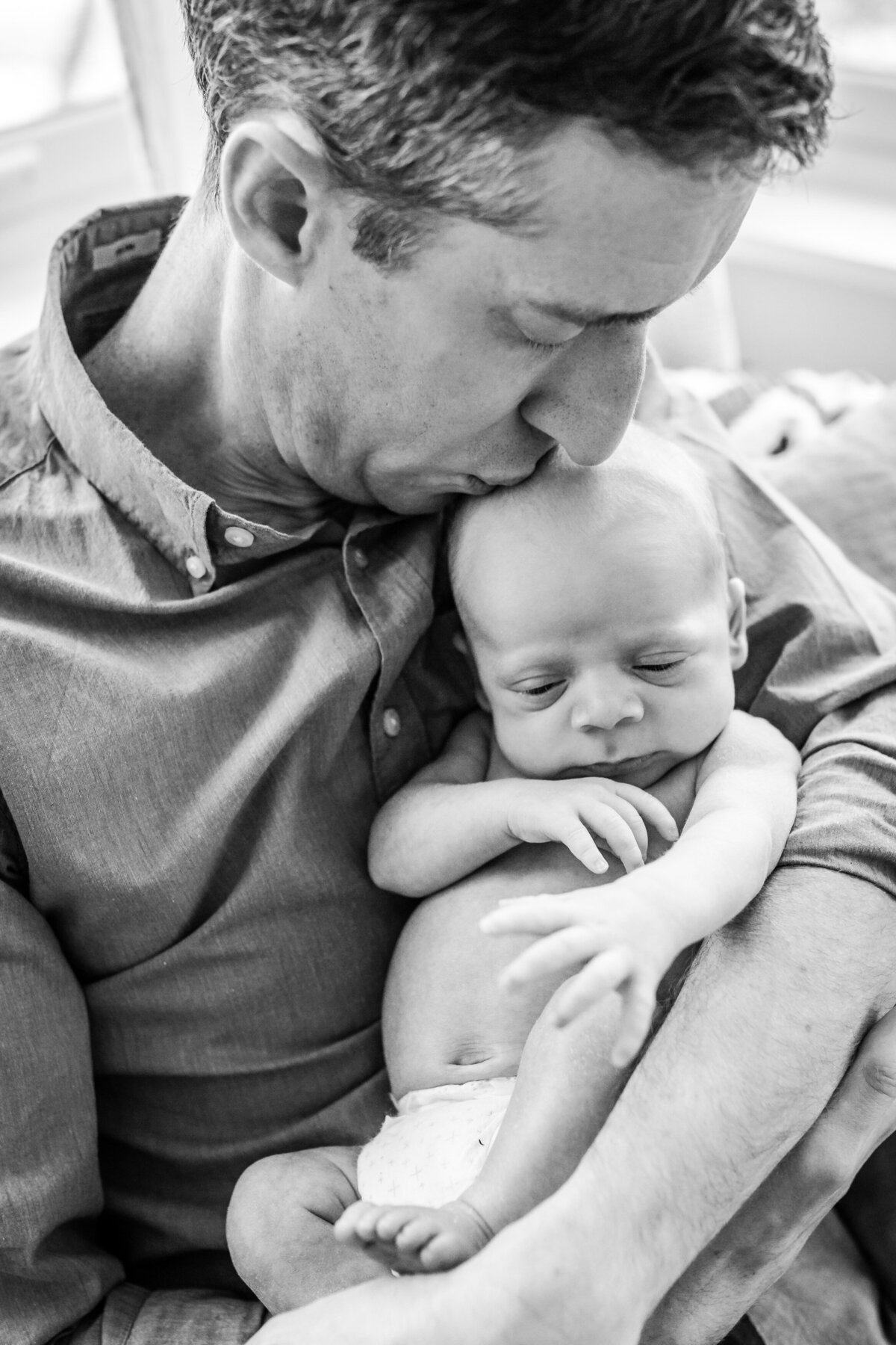 A black and white close-up of a father holding his newborn baby close, gently kissing the baby's head as they both relax.