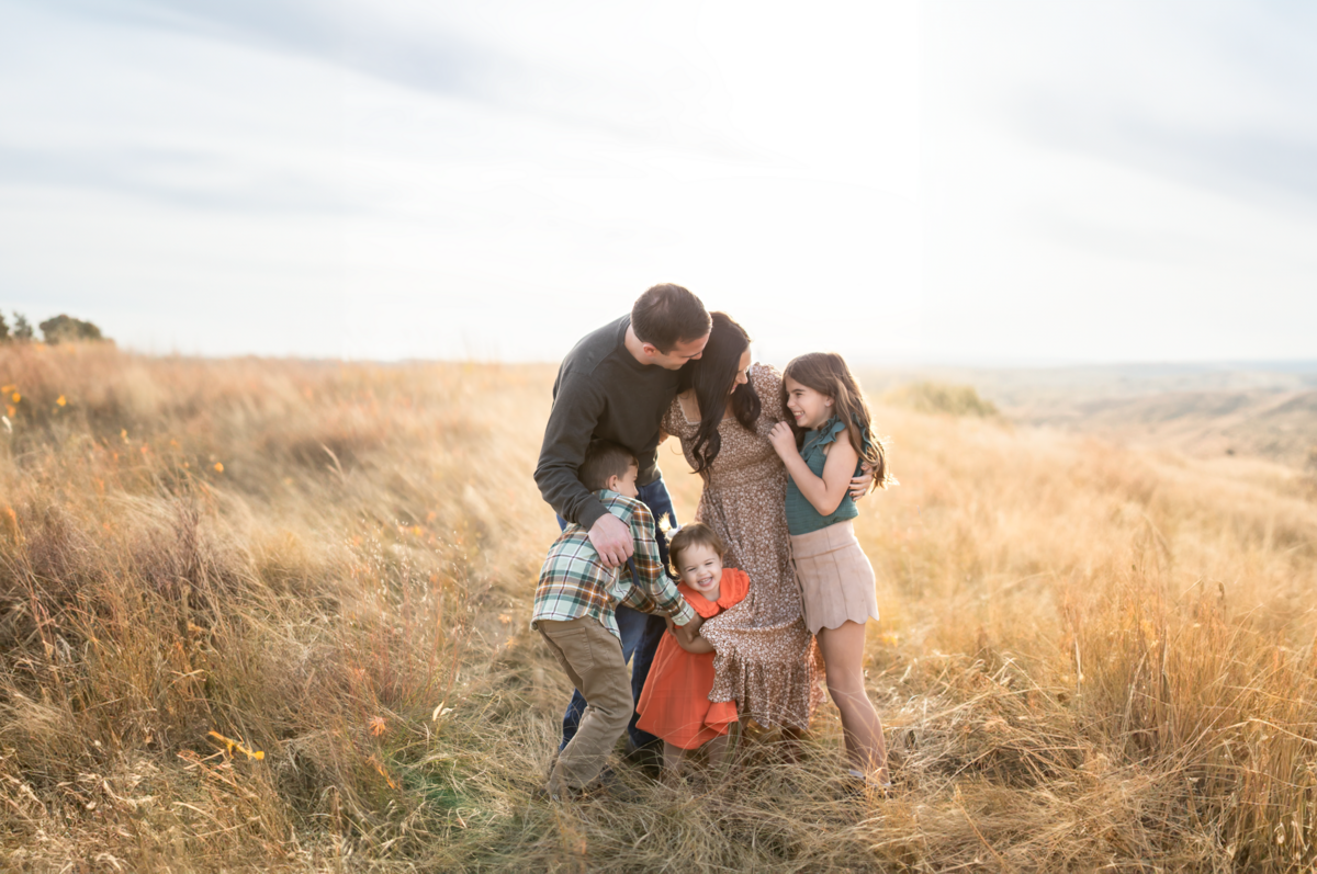a family hugs eachother in a field of golden tall grass at sunset, smiling with eachother in the golden hour sunlight