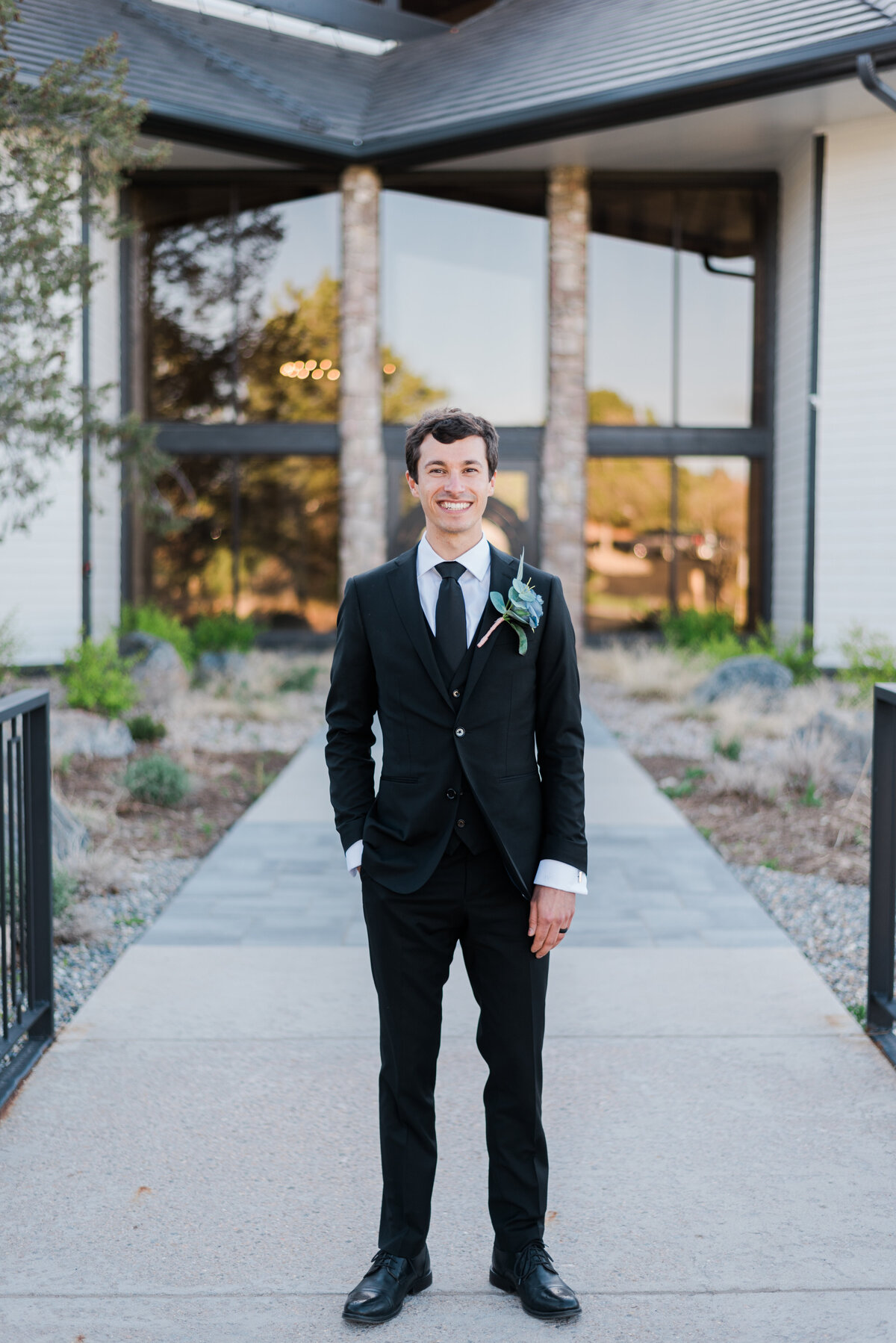 Denver wedding photographer captures the groom in a black suit standing outside The Oaks venue with one hand in his pocket captured by kb radiant photography, denver wedding photographer