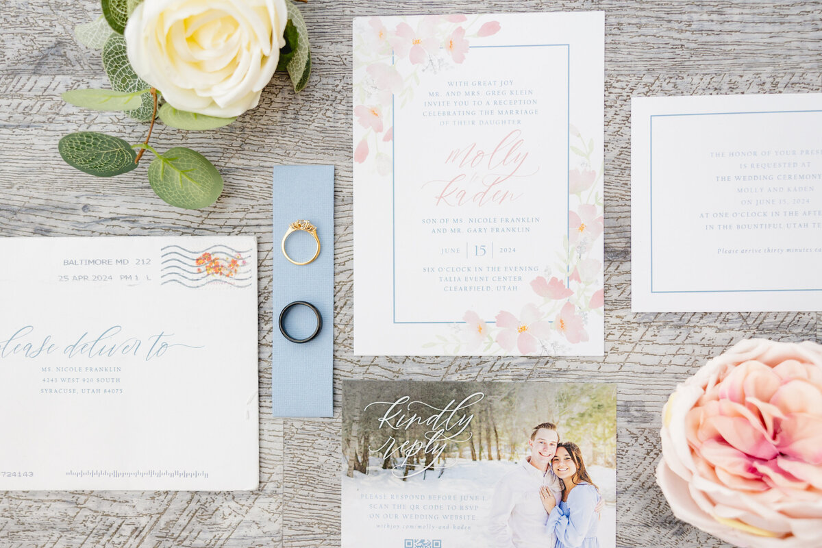 A flat-lay of a wedding invitation set with delicate floral designs, wedding rings, and a white rose, arranged on a rustic wooden surface.