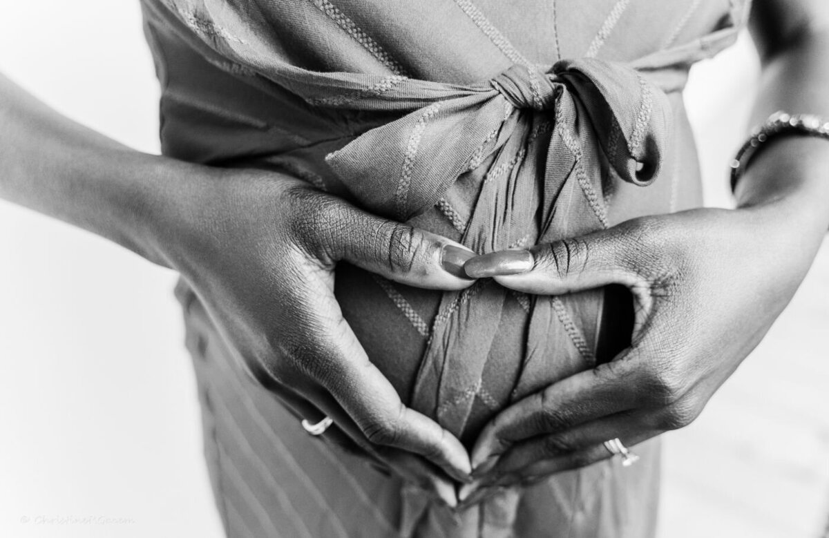  A close-up, black and white portrait of a pregnant woman holding her hands on her baby bump. She is wearing a simple, dark outfit. Her hands are gently placed over her stomach, and the image is focused on her hands and belly, creating a soft, intimate moment.