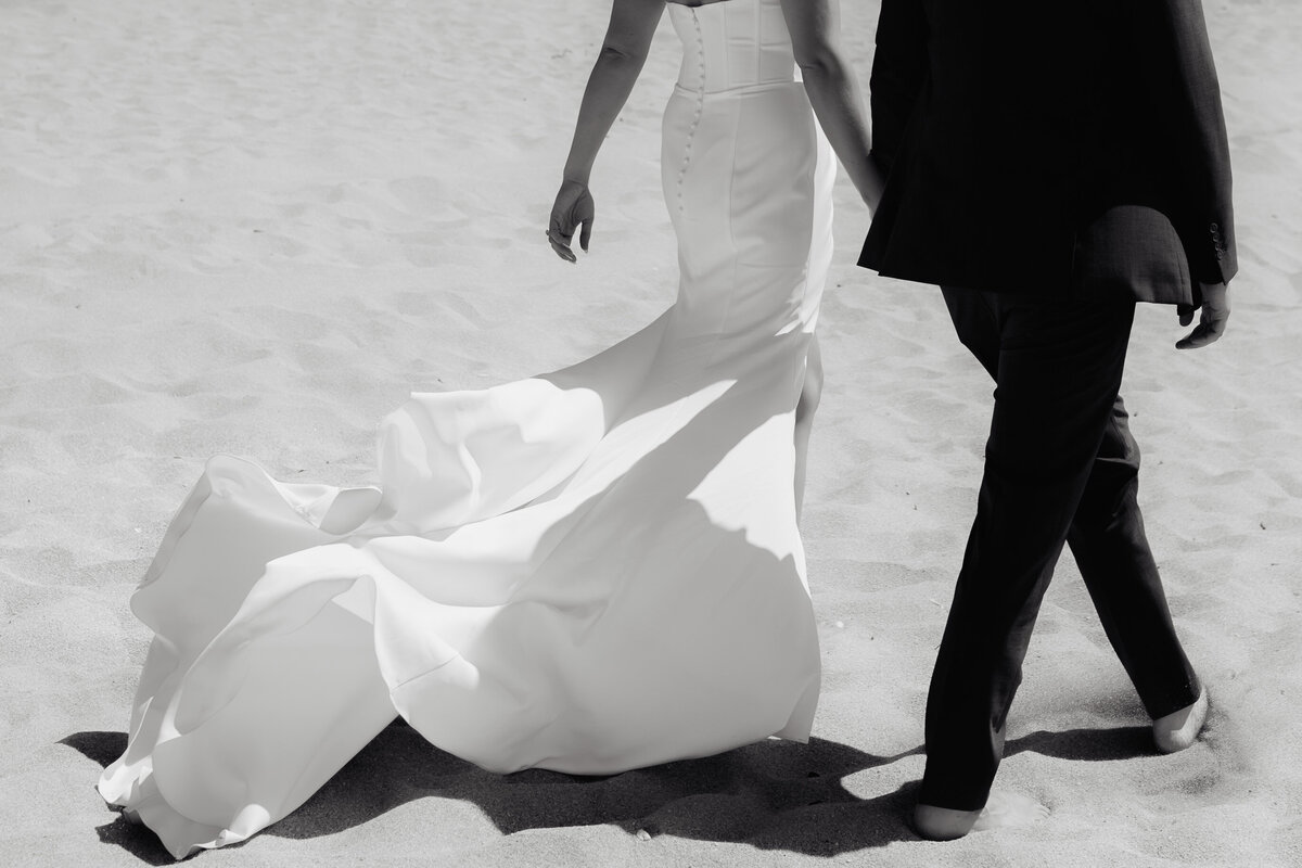 Artistic black-and-white detail of bride’s flowing dress walking barefoot alongside groom on Montara State Beach.