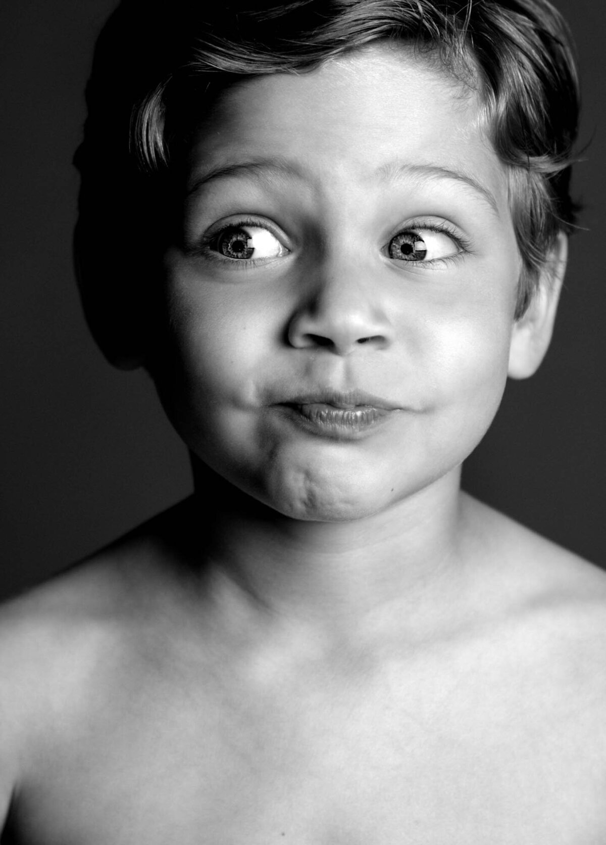 A black and white portrait of a young child with light hair, making a playful, curious expression. The child looks to the side, capturing innocence and wonder.