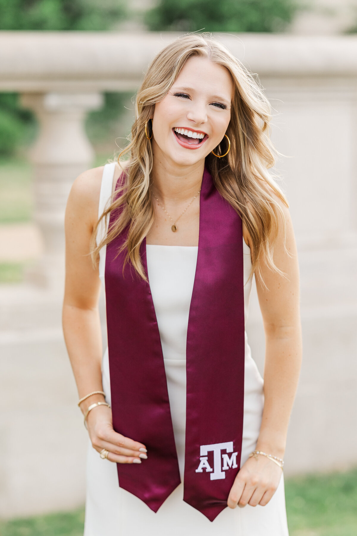 Happy Texas A&M graduate laughing while wearing a maroon stole with the A&M logo over a white dress. She stands outdoors in College Station with natural greenery and stone architecture in the background.