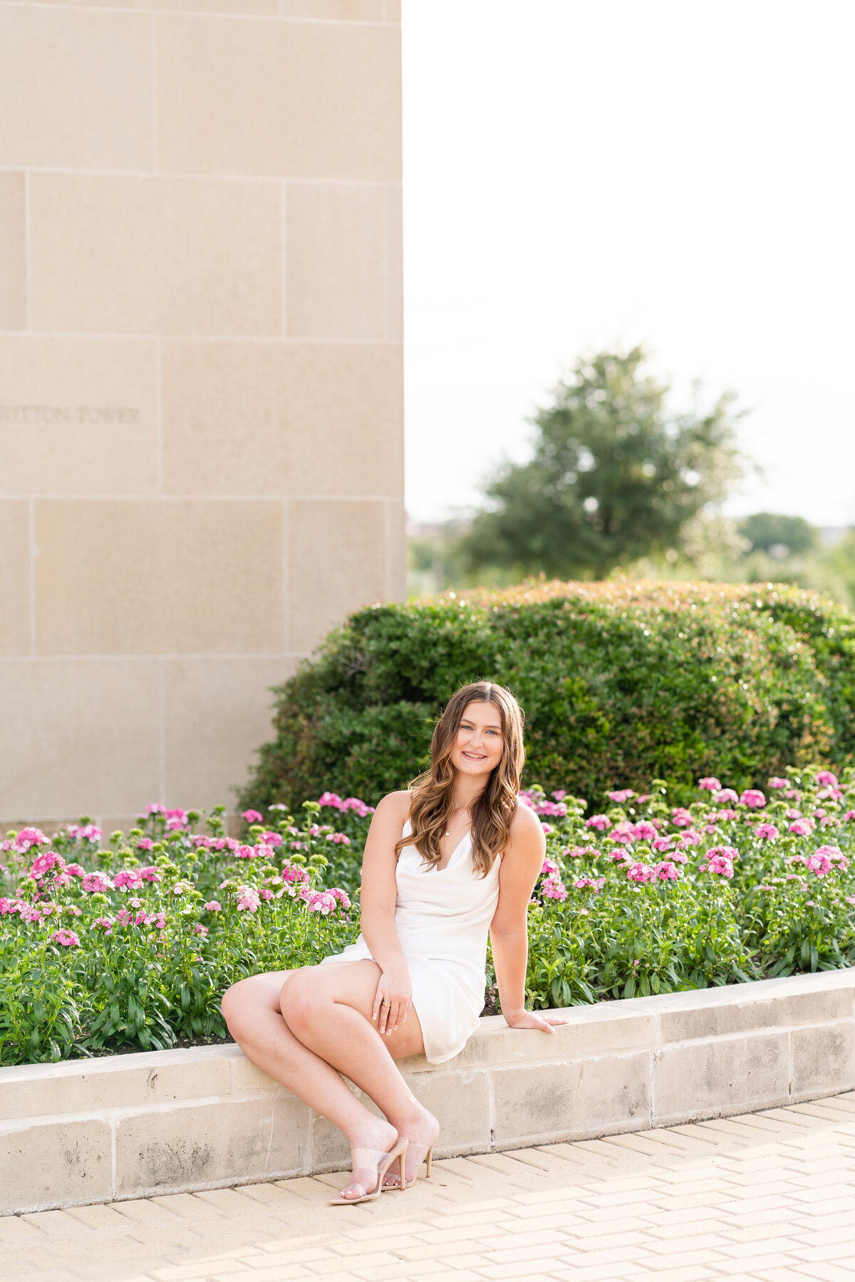 Texas A&M senior girl sitting in white dress in front of colorful flowers at Bell Tower