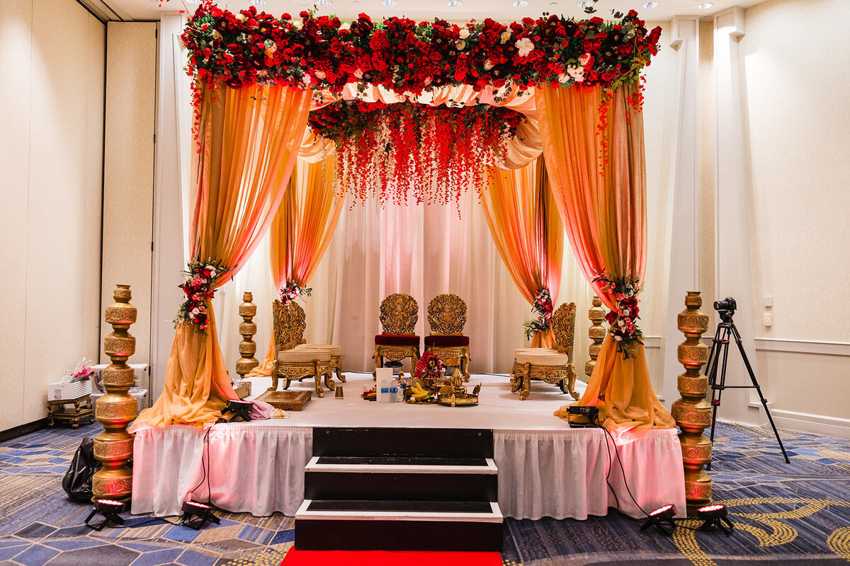 An ornate Indian wedding ceremony stage with red flowers and orange drapes, including a traditional seating area for the bride and groom.
