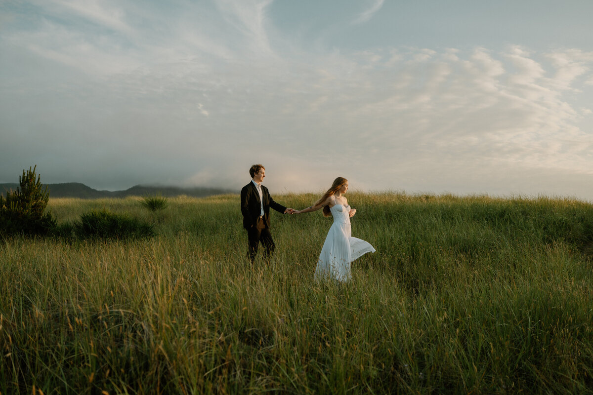 A couple walks through tall green grass at sunset during their Cannon Beach elopement. 