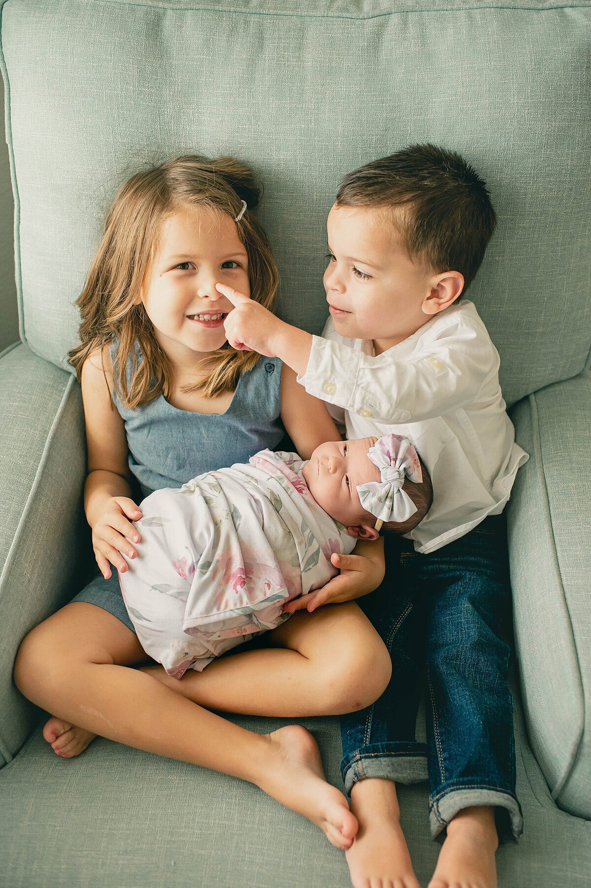 Brother touching sisters nose sitting in chair while she holds new baby sister at their Houston Newborn Photography session