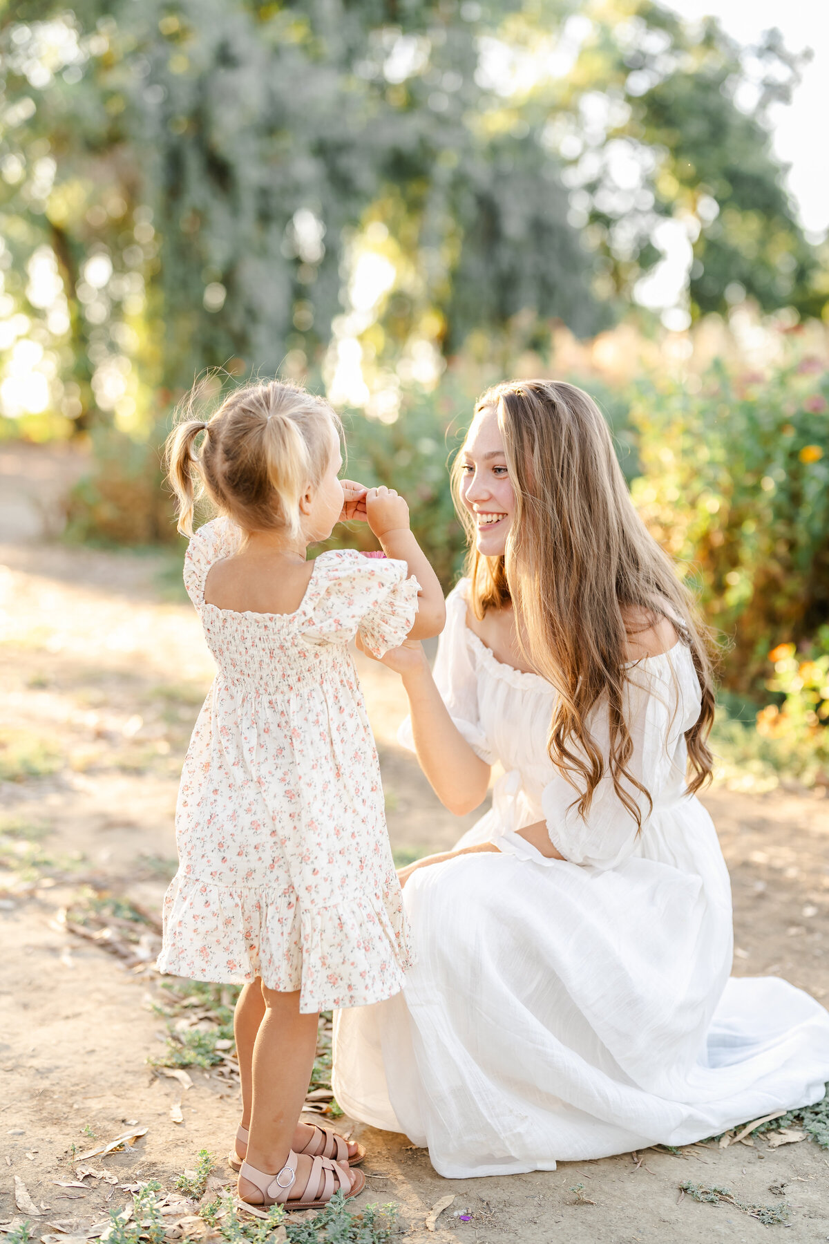 A mother with long blonde curls wearing a white gown kneels and smiles lovingly at her toddler daughter wearing a pale yellow floral dress photographed by Bay Area Family Photographer, Light Livin Photography.