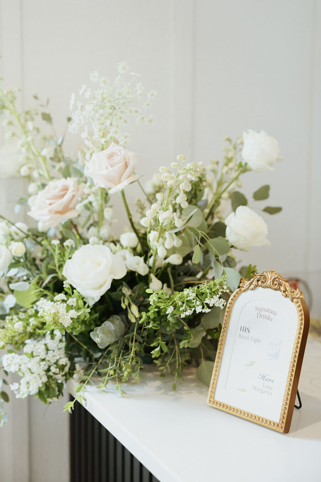 wedding signature drinks sign with his and hers drinks in a gold frame next to florals