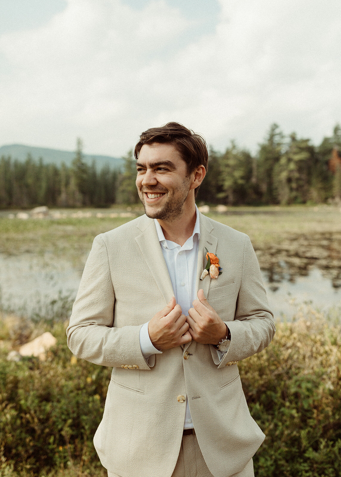 A groom in a beige suit in the Adirondacks with mountains and a lake behind him.