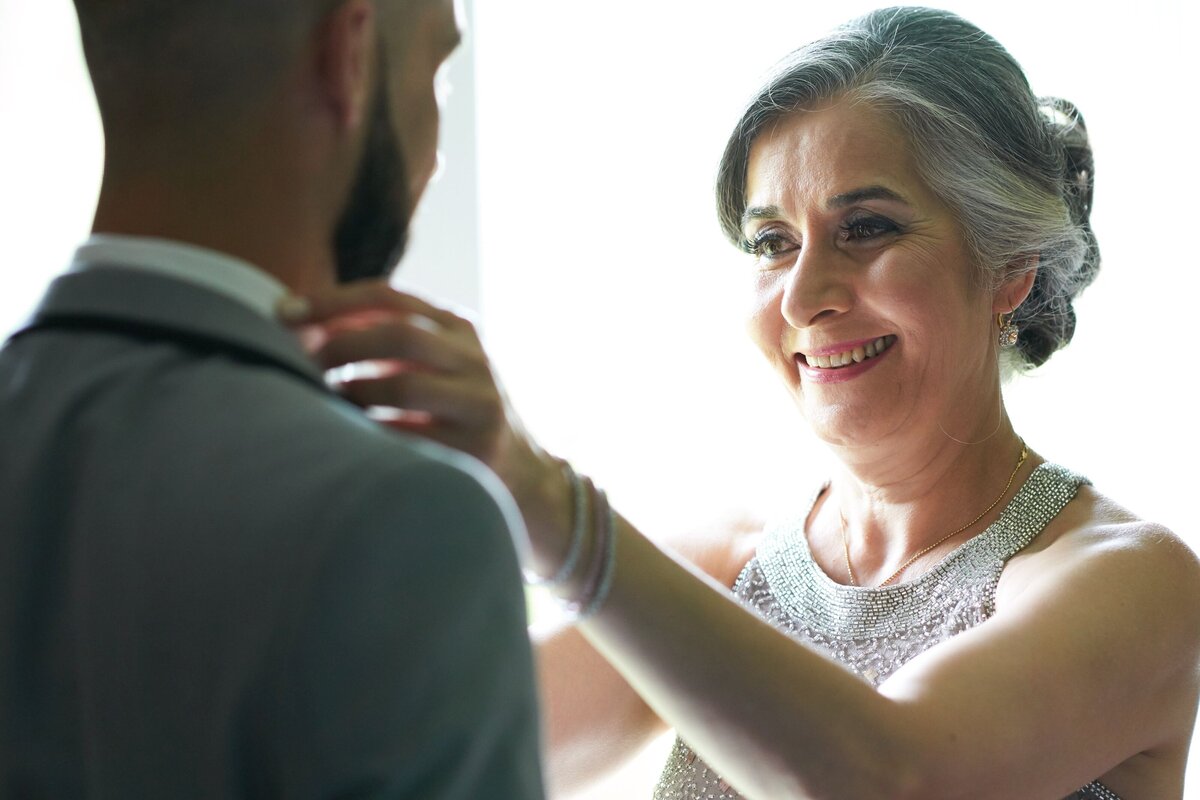 The groom's mother adjusts his collar, adding a tender and supportive touch to his wedding preparations. This image captures a heartfelt moment of maternal care and attention to detail, highlighting the emotional connection and personal support on the wedding day.