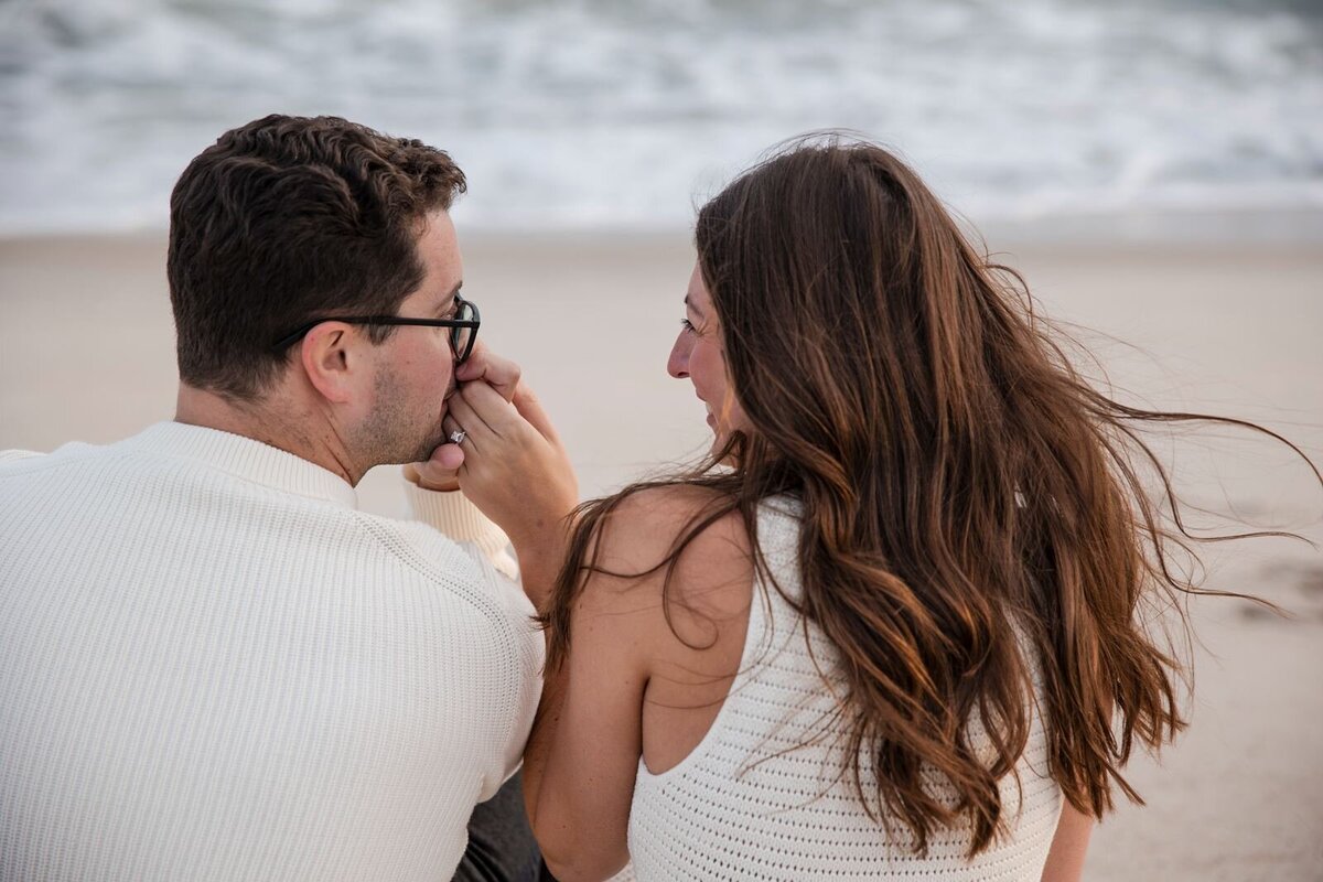 Romantic Fall Hampton's Engagement - kiss on the hand as the couple sits by the beach shore