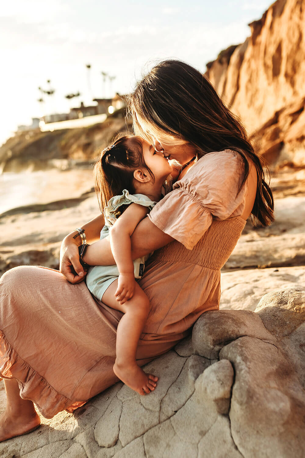 Mom kissing toddler daughter sitting on her lap on a cliff.