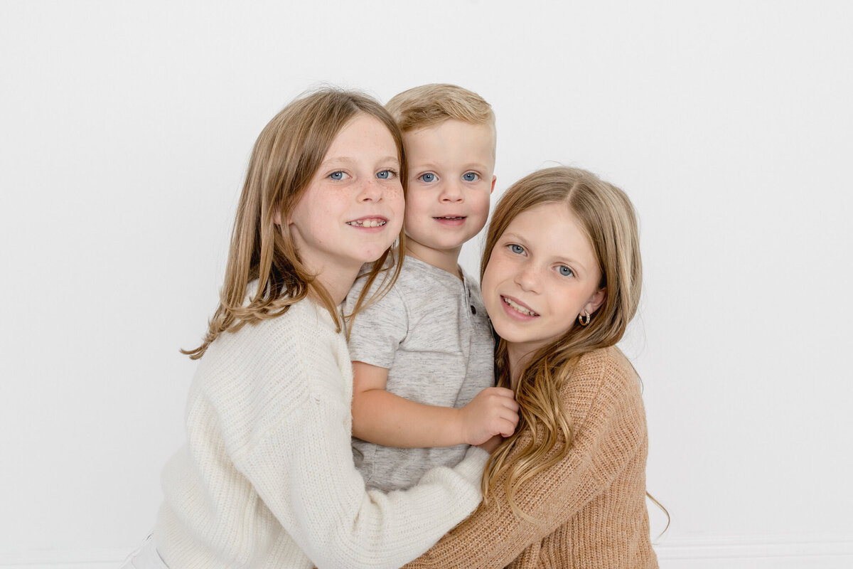 Three young siblings with blonde hair hugging each other with their heads pressed together looking at the camera at a white studio in Tustin.