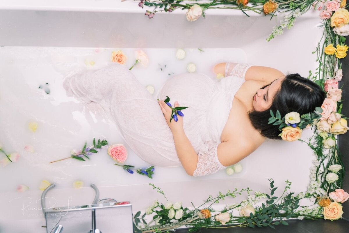 Overhead view of an expectant mother in a floral milk bath holding a small purple flower while cradling her belly in a white lace gown