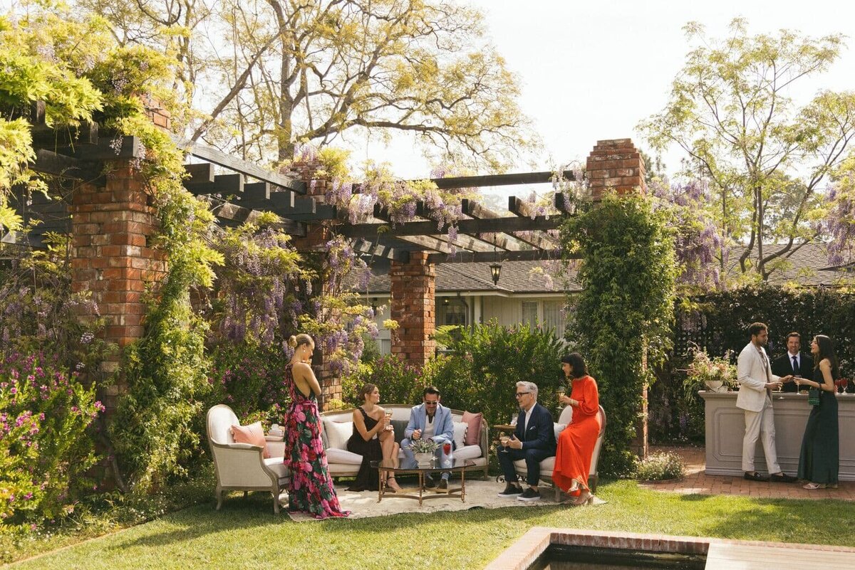 An elegant outdoor garden party with well-dressed guests mingling under a pergola adorned with cascading wisteria