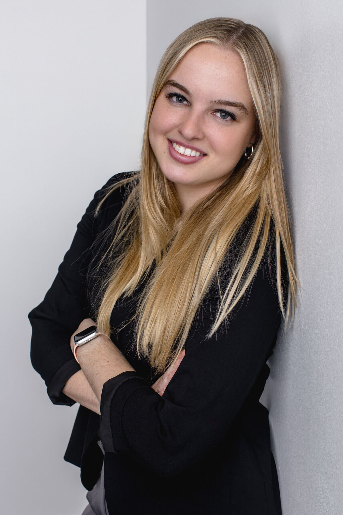 A young woman with long, blonde hair wearing a black blazer leaning against a white wall, smiling at the camera in a white studio.