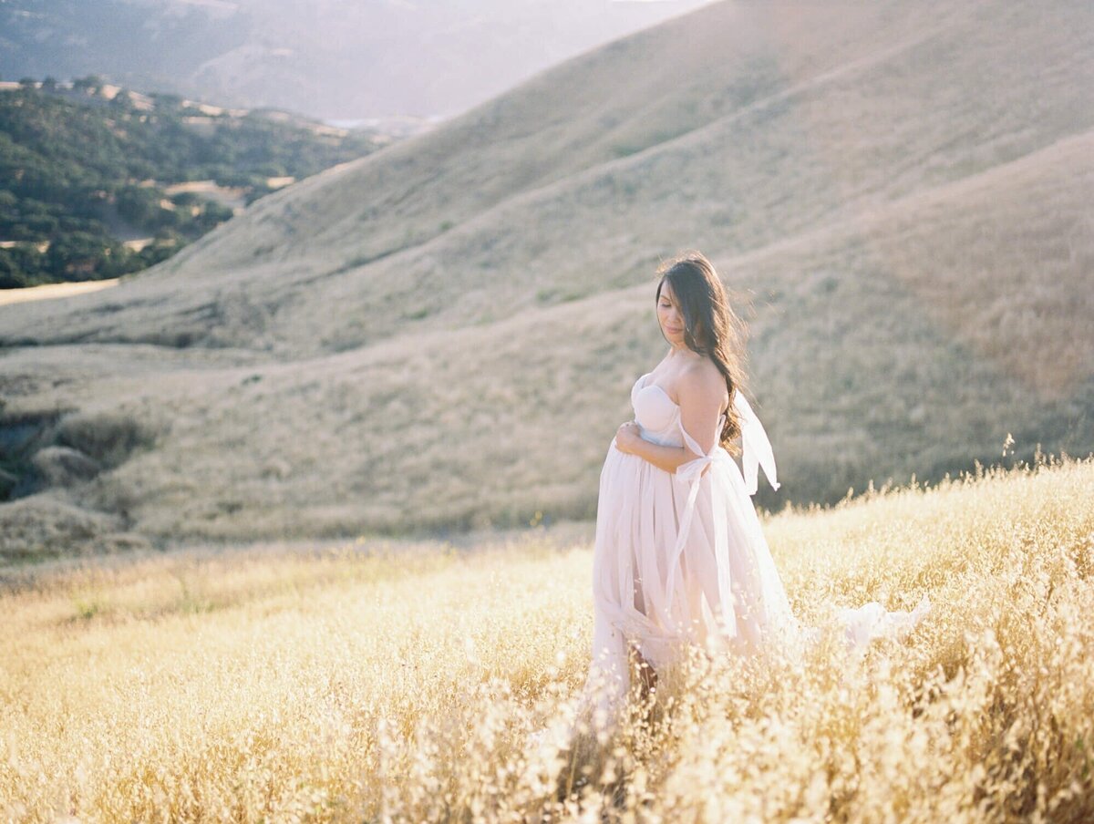 Hillside maternity photoshoot in San Francisco with a pregnant woman standing in a field against hills.