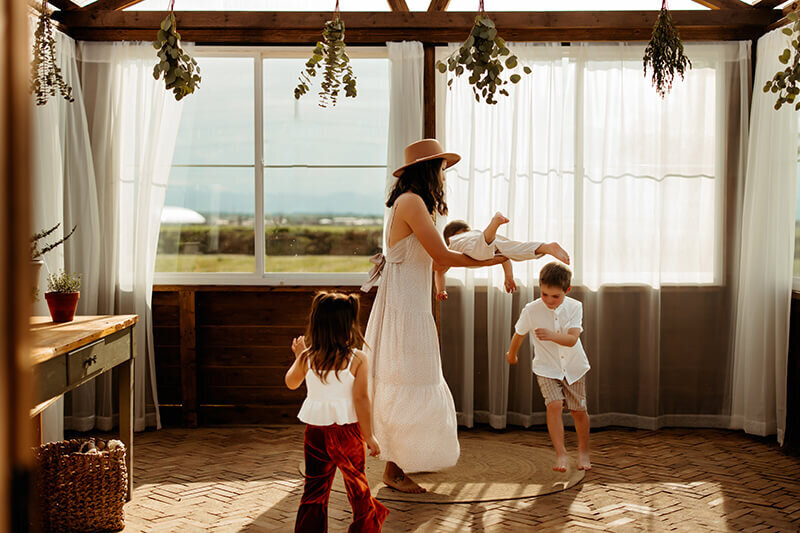 Mom plays with her young kids in a green house while wearing neutral colors on a sunny day