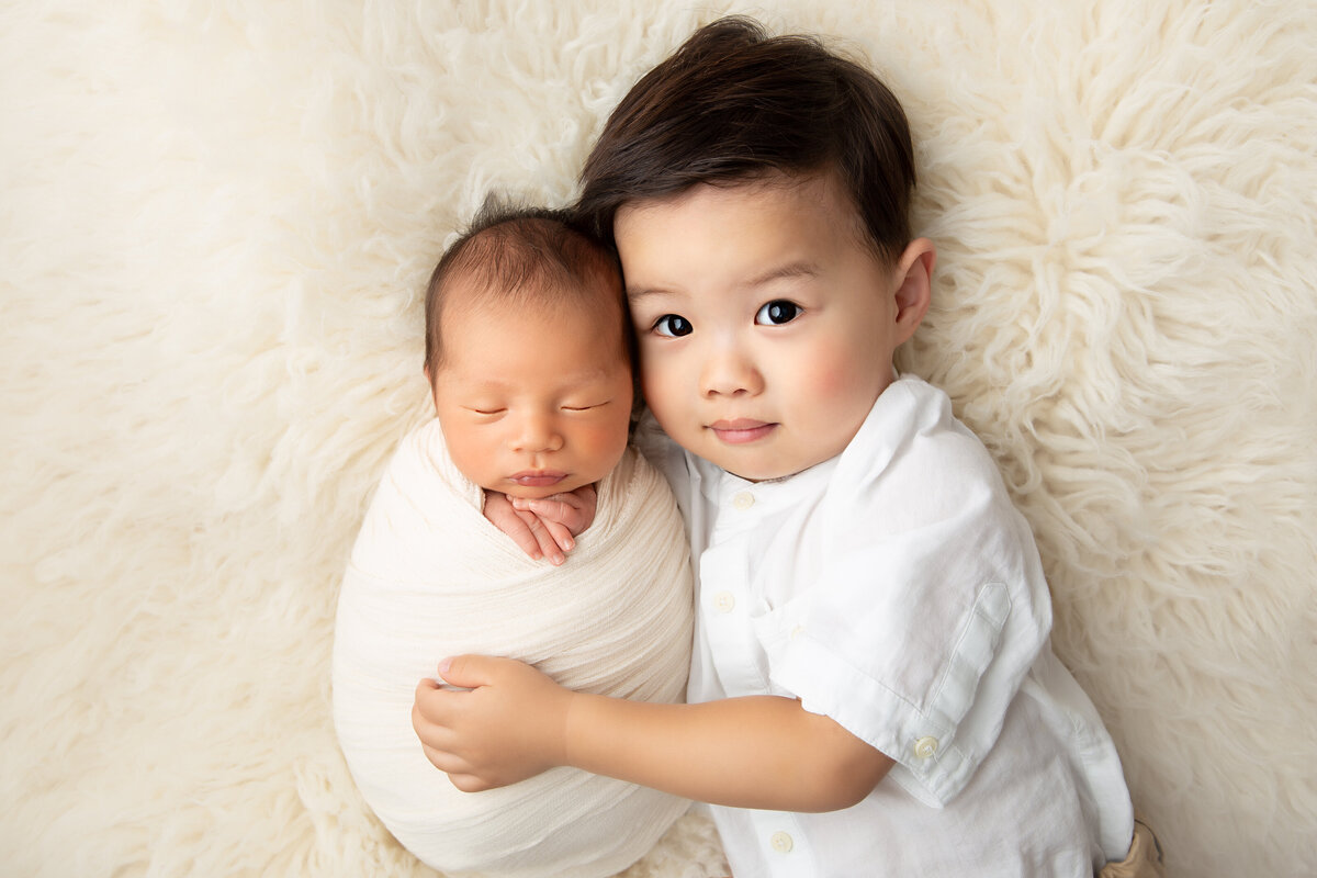 Three year old brother in a white shirt lying next to his wrapped up newborn brother with his hand gently on him.