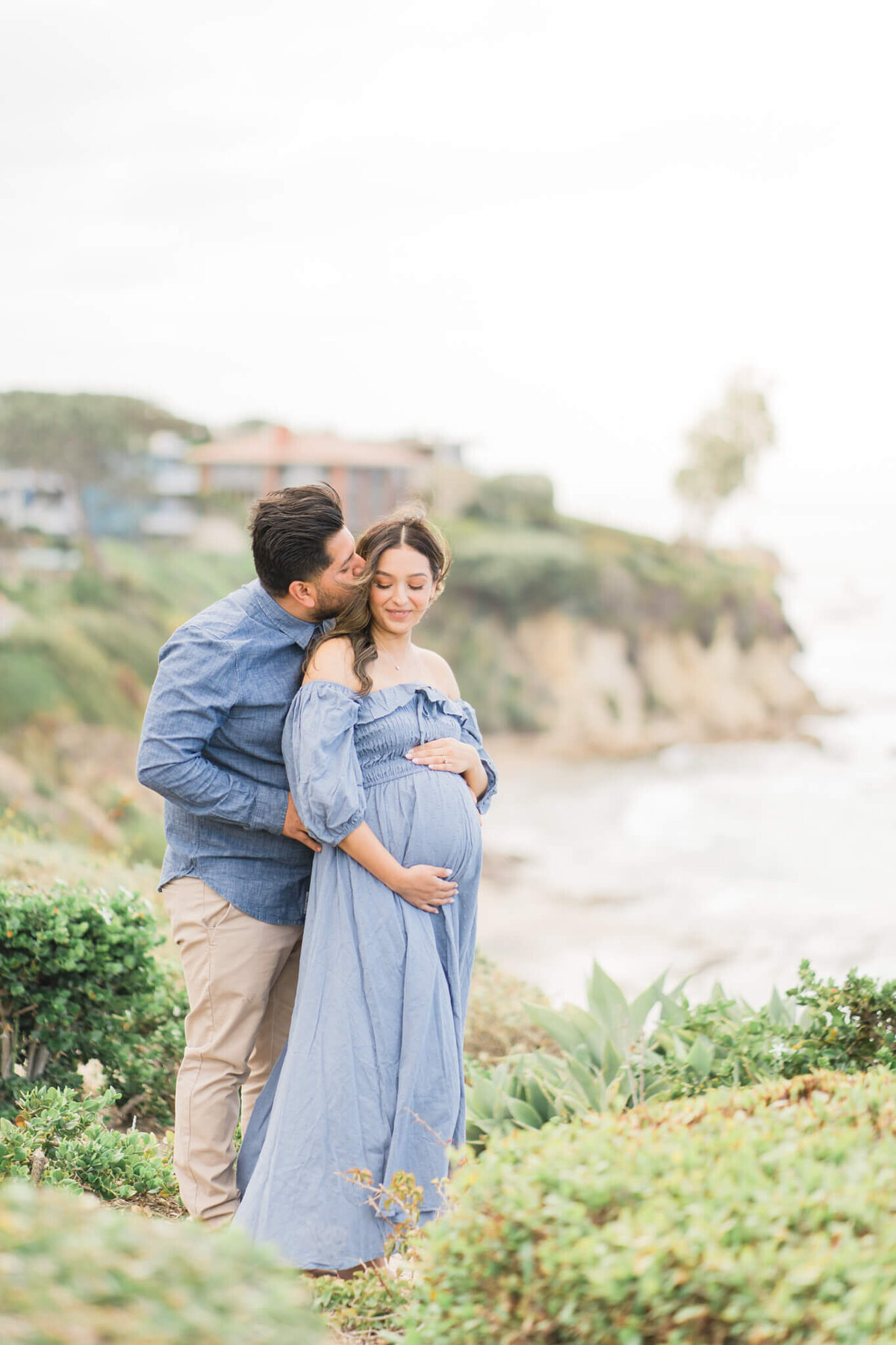 Pregnant couple in light blue outfits standing on cliff by the oceanside in California