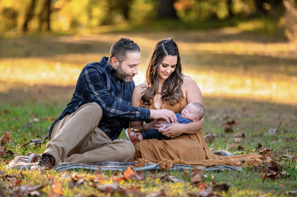 Father and mother looking down at their newborn baby while sitting on a blanket at Sunset in the grass
