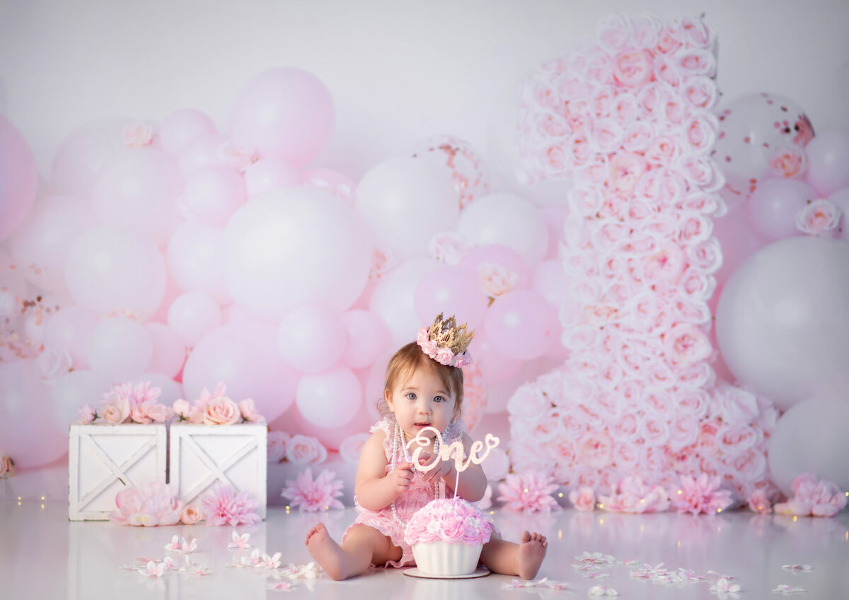 One year old baby girl wearing a crown sitting in front of a beautiful backdrop with pink balloons and florals ready for her cake smash