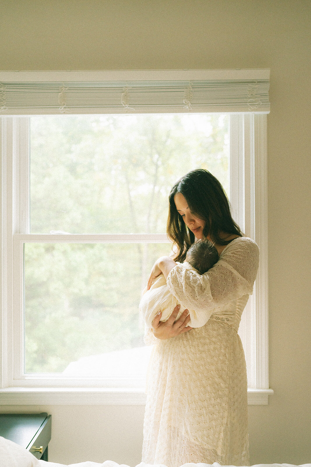 Mother gently holding her newborn baby near a bright window during a lifestyle session at home

