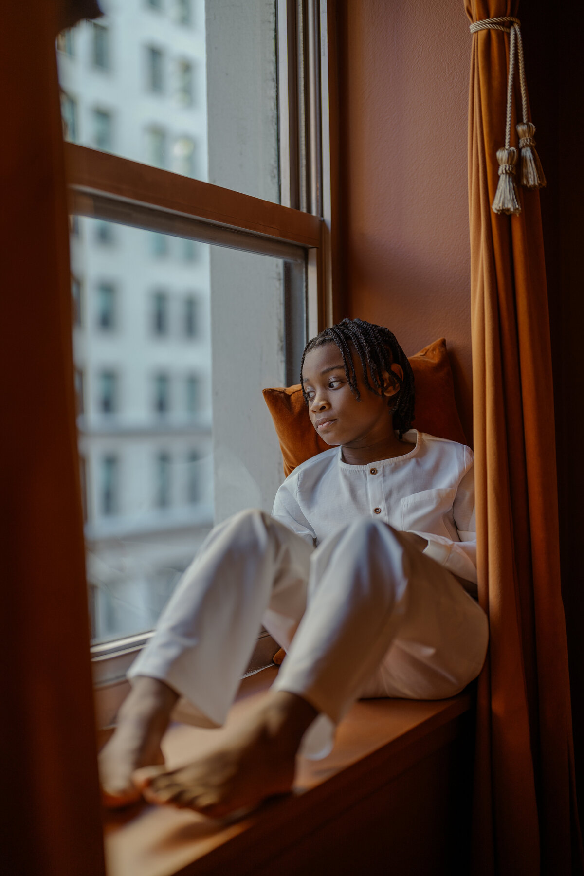 A sophisticated portrait of a young child sitting by a window, gazing thoughtfully outside during a family session in NYC. The warm, natural light and minimalist composition create a timeless legacy-style image.